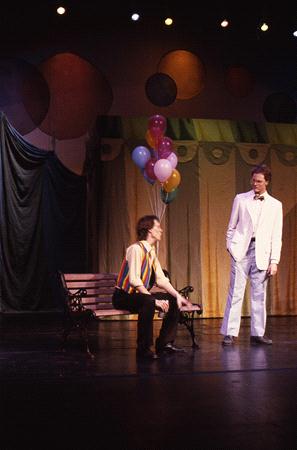 Two actors on a stage. One is sitting on a bench wearing a colorful striped shirt, the other is standing in a white suit. Behind them are multi-colored balloons, and a backdrop with circular patterns and draped fabric.