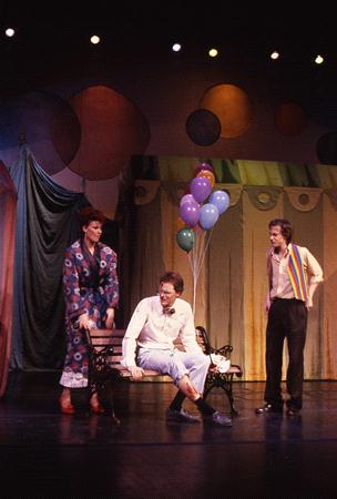 Three men on a stage: one in a colorful robe, another in white sitting on a bench with balloons behind, the third wearing a rainbow-striped sash. The backdrop is a mix of green and warm colors, giving a whimsical atmosphere.