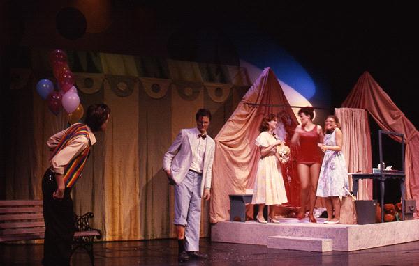 A group of five people is performing on stage with a colorful, tent-like backdrop. One person in a white jacket stands near the center, while others in various costumes are on the right. A bench and balloons are on the left.