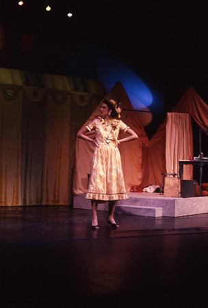 A person in a yellow dress poses confidently on a dimly lit stage. The backdrop features large, draped fabrics in warm colors creating a theatrical set. The lighting casts colorful highlights on the scene.