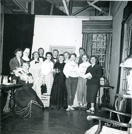 A group of people posing on a stage set, dressed in various costumes. One person is seated holding a bouquet, while others stand around them, smiling. The backdrop includes a window and curtains. A lamp and a table are visible in the foreground.