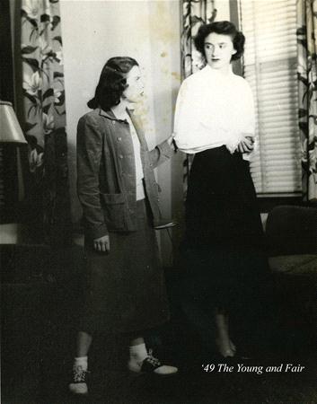 Two women stand indoors. One wears a long skirt and casual jacket, holding the other's arm who is in a white blouse and dark skirt. The room has patterned curtains and a lamp. Text in the corner reads "'49 The Young and Fair.