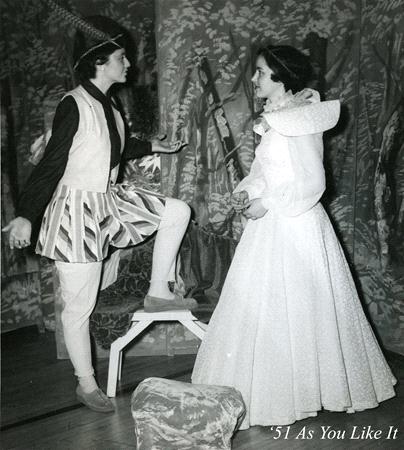 Two actors in a 1951 theater production of "As You Like It." One, in traditional Elizabethan costume, stands with one foot on a stool. The other, in a long gown with a large collar, faces them. The background features nature-themed scenery.