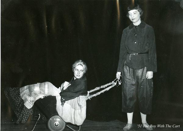 Two people on stage performing a play. One person is sitting in a small cart, wrapped in a blanket, while the other stands, pulling the cart with ropes. The backdrop is dark, and the scene is labeled "51 The Boy With The Cart.