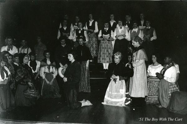 A black and white photo of a group of people in period costumes on stage. Some are kneeling while others stand behind them. The backdrop is dark, and the setting appears to be a theatrical performance titled "The Boy With the Cart.