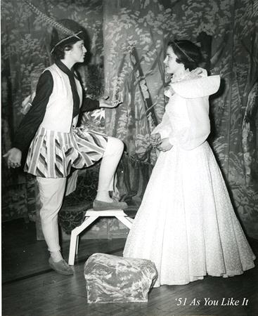 Two actors in vintage costumes perform a scene from "As You Like It." One is in a patterned outfit with a hat, standing on a step; the other wears a long, elegant gown. Text reads: "'51 As You Like It." The backdrop features a forest design.