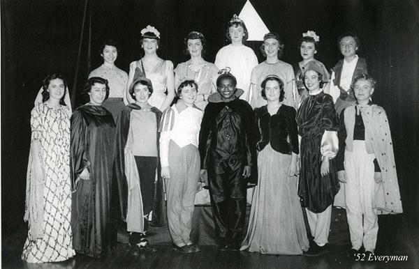 A black and white photo of thirteen individuals dressed in various theatrical costumes, standing on a stage. They are arranged in three rows, smiling at the camera. A dark curtain serves as the backdrop.