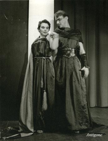 Two actors on stage in dramatic costumes. The person on the left wears a long dress with a cloak, and the person on the right wears a striped robe with armor-like accessories. The backdrop is simple, and "Antigone" is written in the corner.