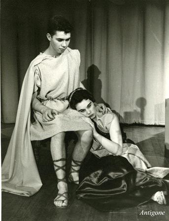 A black-and-white photo shows two actors on stage. One is seated, wearing a tunic and cloak, with an arm draped over the other, who is kneeling and resting their head on their lap. The caption reads "Antigone" in the corner.