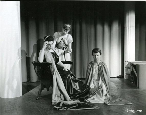 Black and white photo of three people in theatrical costumes on stage. One sits in a chair with a cape, one stands holding a staff, and one kneels with a cloak. Heavy drapery and columns form the backdrop. The word "Antigone" is in the corner.