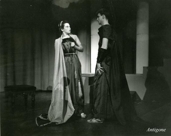 Two actors in classical Greek costumes perform a scene on stage. The woman wears a gown and a cape, holding an object, while the man stands opposite her in a tunic and cloak. The stage has pillars in the background. "Antigone" is written in the corner.