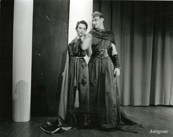 Two people in Greek-style costumes stand on a stage. The woman on the left wears a long dress, and the man on the right is in a tunic with a cape and arm guards. The setting suggests a theatrical performance of "Antigone.