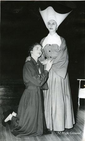 Two women in theatrical costumes are posing on stage. One is kneeling in a dark dress, while the other stands in a flowing gown with a large white headdress. The scene is dramatic, set against a dark background. Text reads "'52 High Ground.