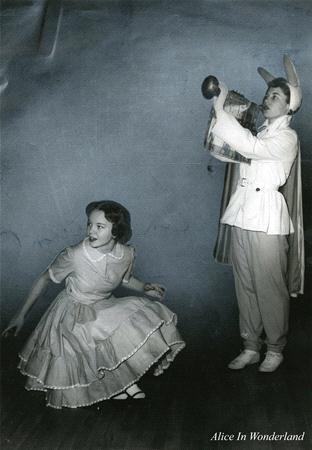 A person dressed as Alice in a light-colored dress crouches slightly, looking intrigued. Another person, dressed as a rabbit with large ears and a trumpet, stands beside them. The image has a vintage look. Text on the image reads "Alice In Wonderland.