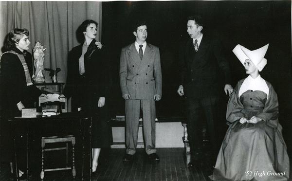 A black and white photo of four standing people engaged in a scene and one seated person wearing a nun's habit with a large white headpiece. The room includes a table with a statue and a chair.