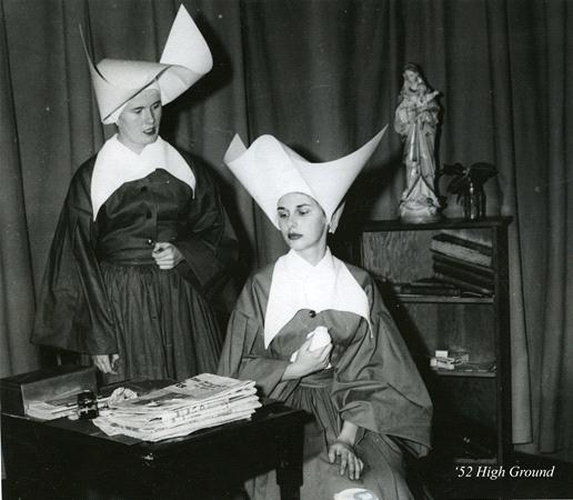 Two women wearing traditional nun habits with distinctive wide headpieces. One is seated holding a small item, the other stands nearby. A statue and books are on a shelf behind them. The setting appears to be indoor with curtains in the background.