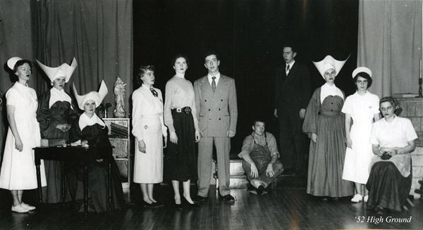 A group of people pose on stage in a theatrical setting. Some wear nun costumes with distinctive headpieces, while others wear mid-20th century attire. A few are seated, and a man stands in the background. The stage is dimly lit.