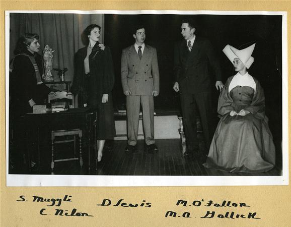 Five people performing on stage with a vintage setting. Four are standing and dressed in formal attire, while one is seated in a traditional nun's habit. Props and a statue are on the table to the left. Names are handwritten below the photo.