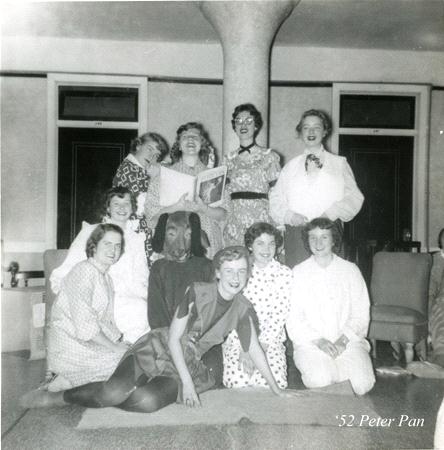 A group of women in 1950s-style clothing pose indoors. Most are seated, with a few standing behind. One holds a book, and a person in a dog costume is in the center. A woman in front wears a Peter Pan costume. Text reads "'52 Peter Pan.