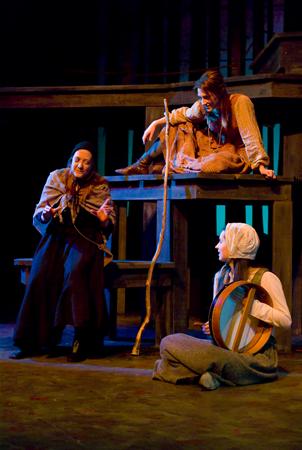 Three actors perform on stage in a dimly lit theater. One actor sits with a staff, another dressed in medieval attire is seated on a platform, and the third, in a bonnet, holds a round object. The scene suggests a rustic or historical setting.