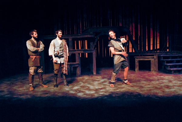 Three actors in period costumes perform on a dimly lit stage with a rustic set. One actor stands to the right in a dynamic pose, while the other two watch from the left, engaged in the scene.