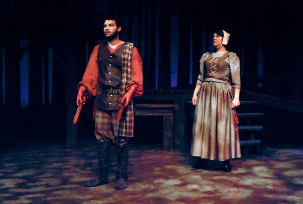 A man and woman in historical costumes stand on a dimly lit stage. The man wears a tunic and plaid trousers, holding a dagger, while the woman wears a long dress and bonnet. The background is dark with wooden structures.