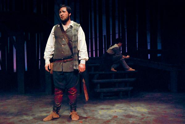 A man stands on stage in medieval attire, wearing a brown tunic, red trousers, and brown shoes. Behind him, another person sits on steps in the background, partially obscured by shadows. The setting has a dark, wooden backdrop.