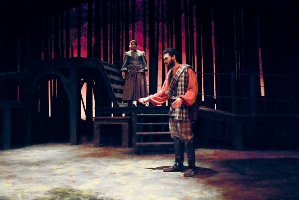 Two actors perform on a dimly lit stage with a forest backdrop. One stands on a higher platform, dressed in medieval-style clothing, while the other stands below, gesturing passionately. The scene evokes a dramatic, historical atmosphere.