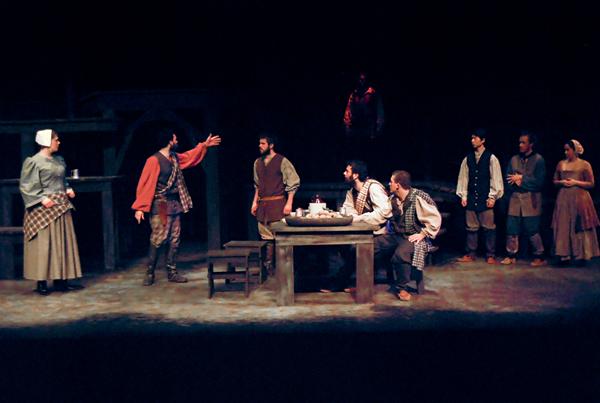 A theatrical scene set in a dimly lit room with actors in period costumes. One person stands gesturing passionately while others sit and listen around a wooden table. A few people stand in the background observing the interaction.