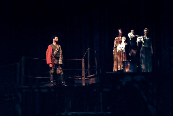 A dimly lit stage scene with a man in red and plaid standing on the left. On the right, a group of people holding dolls stands slightly elevated. The atmosphere is dark and dramatic.