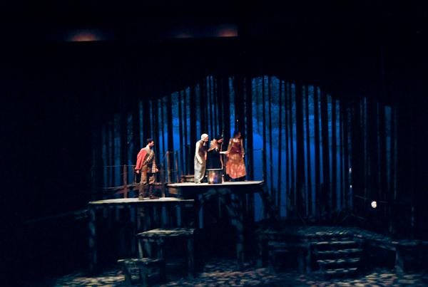 A dimly lit stage with four performers dressed in period costumes, standing on a platform surrounded by a backdrop resembling tall, dark trees. The blue lighting creates a mysterious atmosphere.