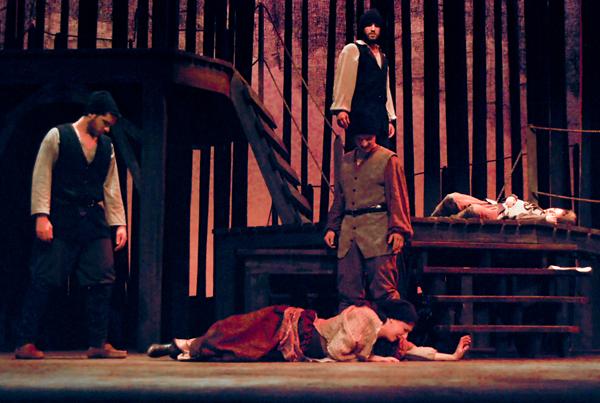 A theatrical scene with actors in historical costumes. One actor lies on the floor while others stand around, and another is positioned dramatically on a structure with wooden bars. The lighting is dim and moody.