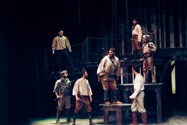 A group of actors on stage in a dimly lit theater perform a fantasy scene. They are dressed in medieval-style costumes, including loose shirts and boots, standing on wooden platforms and stairs, with some holding props like staffs or bows.