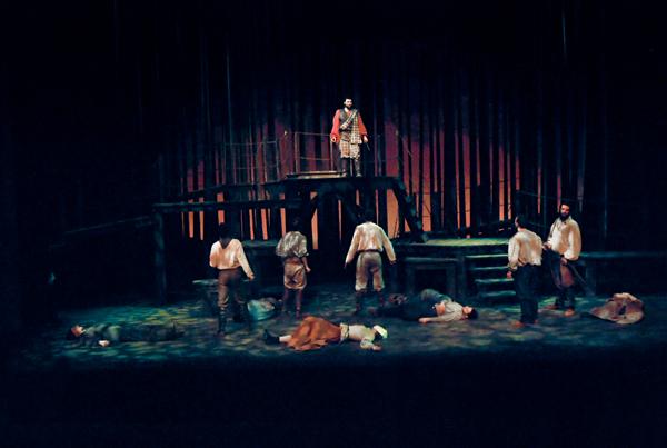 A theatrical stage scene depicting several people fainted or lying down, while a person stands elevated, holding a sword or staff, and observing the scene. The backdrop is dark with a wooden structure and dim lighting, creating a dramatic atmosphere.