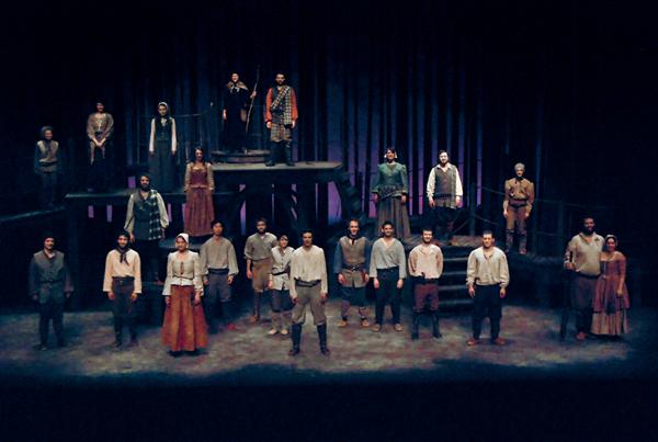 A group of performers stands in a theater setting. They are arranged on two levels, with some on a raised platform. The background is dark, with subtle lighting highlighting the cast. They wear period costumes, such as tunics, skirts, and boots.