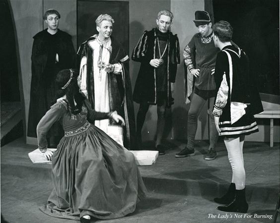 A black and white stage scene from a medieval play with six actors in period costumes. A seated woman in a long dress gestures dramatically as five men in robes and tunics stand around her, engaged in animated conversation.