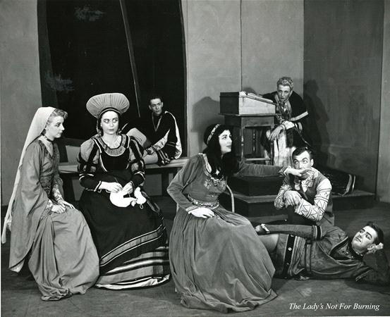 A black-and-white scene in a medieval-themed play depicts six actors in period costumes. One woman is seated, wearing a tall headdress. Two others kneel nearby. A man lies on the floor, and two more are in the background, one by a harpsichord.