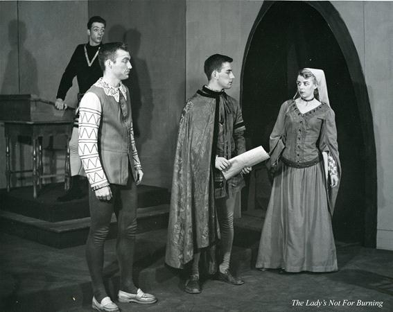 A black and white scene from a play with four actors in medieval costumes. One stands on a raised platform, while two others converse, one holding a scroll. A woman in a long dress and headpiece stands nearby. Text at the bottom reads "The Lady's Not For Burning.