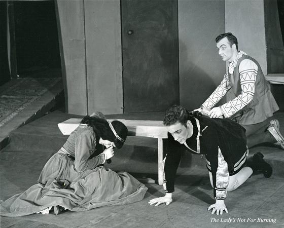 Three actors perform a scene from "The Lady's Not for Burning." Two men in period costumes kneel by a bench on stage, one with his hand on the ground and the other looking up. A woman in a long dress sits on the ground, resting her head on the bench.