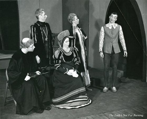 A black and white stage photo shows five actors in period costumes. Four sit on a bench, while one stands, facing them. The setting suggests a medieval play, noted as "The Lady's Not For Burning" in the corner.