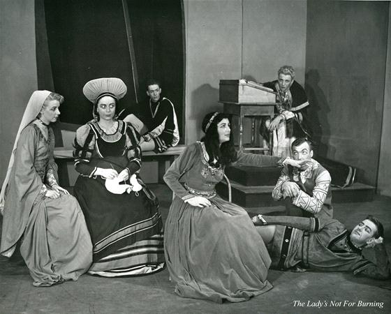 A theatrical scene with actors in medieval costumes on stage. A woman sits with hands folded while another stands beside her. Three men are seated in the background, and a man lies on the floor, interacting with a man crouched next to him.