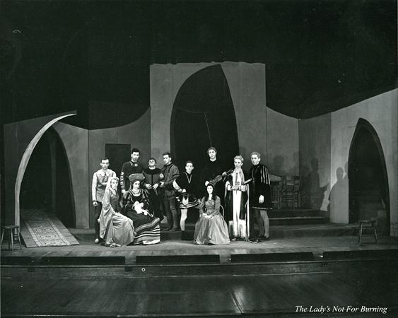 A black and white photo of a theater stage with a group of actors in period costumes posing. The backdrop features abstract arches and curves. Some actors are standing, while others are seated. The play title "The Lady's Not For Burning" is visible.