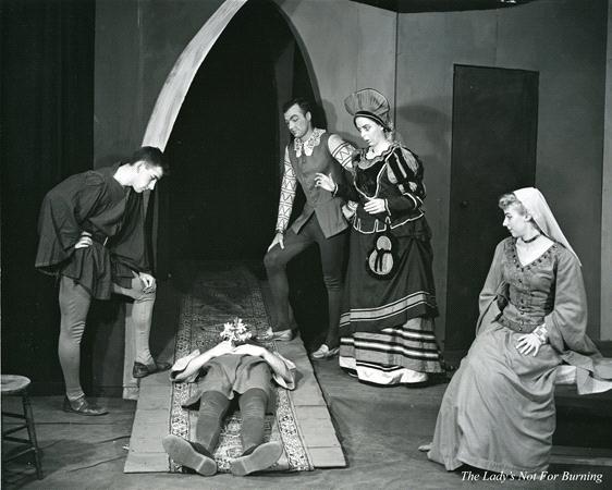 A black and white photo of five actors performing a scene from a play on stage. Four actors stand or kneel around another actor lying on the floor. The scene appears historical, with costumes reflecting an earlier period. The stage backdrop includes an arch.