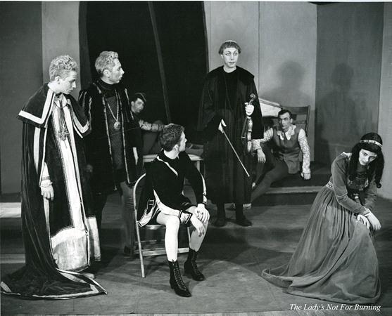 A black and white photo of a theatrical scene. Six actors in medieval costumes are on stage with a backdrop of arches. One actress is kneeling in a long dress, looking distressed. The title "The Lady's Not For Burning" is visible in the lower right corner.