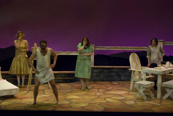 A theatrical scene with four actors on stage. One man is in front, wearing a tunic, making an expressive pose. Three women stand behind him, dressed in period costumes. The set includes a table with chairs and a painted backdrop.