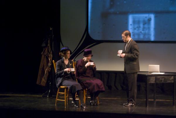 Three actors in period costumes perform on stage. Two women sit and drink from teacups, while a man stands nearby holding another cup and saucer. A black-and-white image is projected in the background.