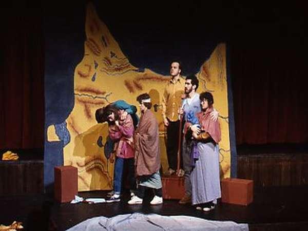 A group of actors on stage perform in front of a large, colorful abstract backdrop. They wear a variety of costumes, standing and interacting with each other. The scene is theatrically lit, suggesting a dramatic or comedic moment.