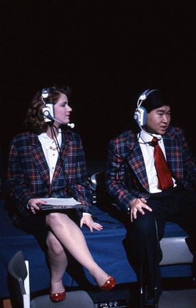 Two people wearing plaid blazers and headsets sit on a table. The woman on the left holds a clipboard, while the man on the right appears to be speaking. They are in a dimly lit setting, perhaps at an event or broadcast.