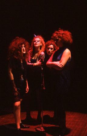 Four people with wild, curly hair sing into a single microphone on a dimly lit stage, creating an intense and dramatic atmosphere.