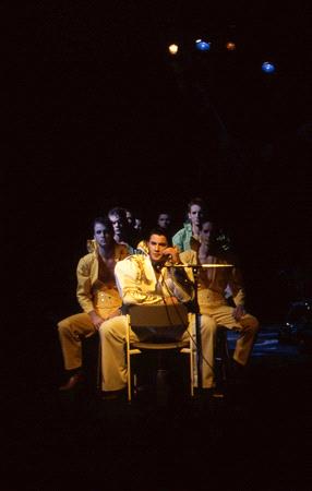 A performer in a white outfit sits on a chair, holding a microphone. Behind him, four people in matching yellow outfits sit closely together on a dark stage. The spotlight is on the performer, while the background remains dimly lit.